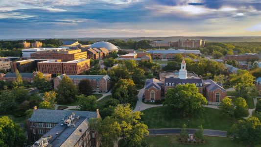 Aerial view of a university campus with brick buildings, green lawns, and a white clock-tower building at the center during late afternoon sun.
