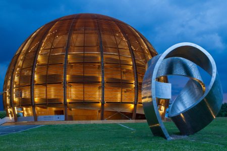 Large copper dome sculpture with glowing interior lights, surrounded by a twisting silver ribbon sculpture at dusk.