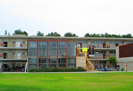 Two-story campus building with a glass central atrium, yellow exterior stairs on the right, people on balconies, and a green lawn in front.