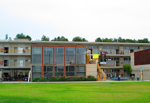 Two-story campus building with a glass central atrium, yellow exterior stairs on the right, people on balconies, and a green lawn in front.