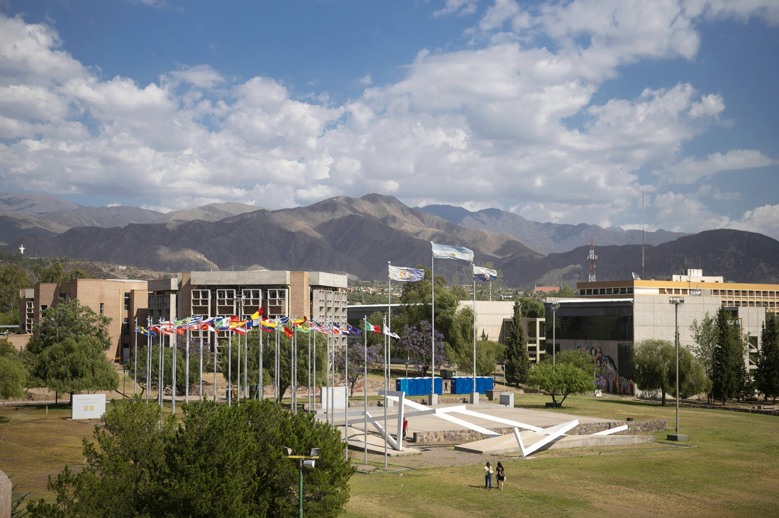 University campus with colorful flags on flagpoles, modern buildings, and a mountain range in the background under a partly cloudy sky.