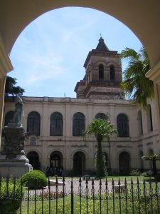 Courtyard of a historic building with arches, a statue on a pedestal, and palm trees framed by a wrought-iron fence.