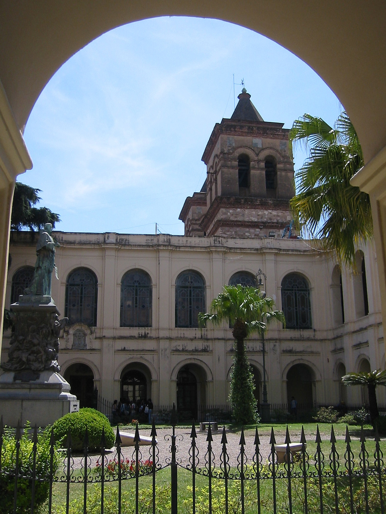 Courtyard of a historic building with arches, a statue on a pedestal, and palm trees framed by a wrought-iron fence.