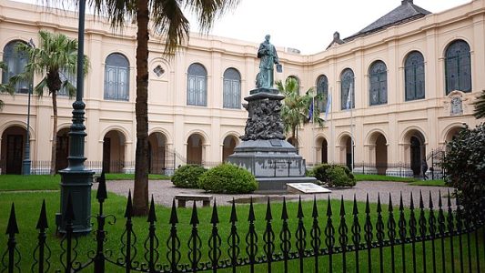 Statue on a pedestal in a courtyard with arches, palm trees, and a decorative iron fence in the foreground.
