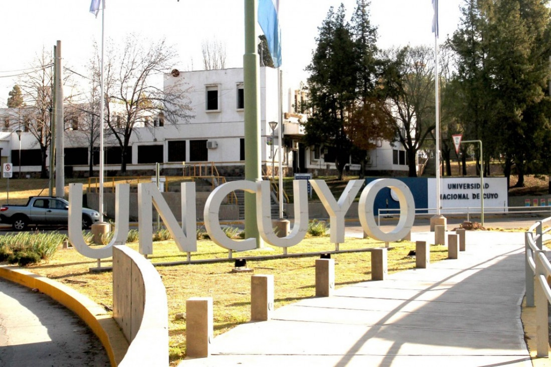 Large white UNCUYO letters on a grassy plaza at a university campus, with a walkway and buildings in the background.